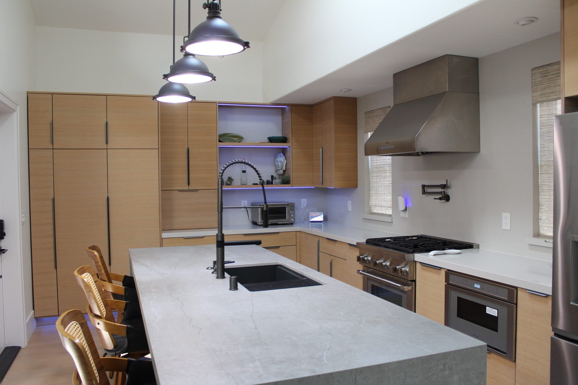 Full kitchen reveal with rift oak cabinetry, waterfall-edge quartz island, stainless range hood, and cane-back barstools