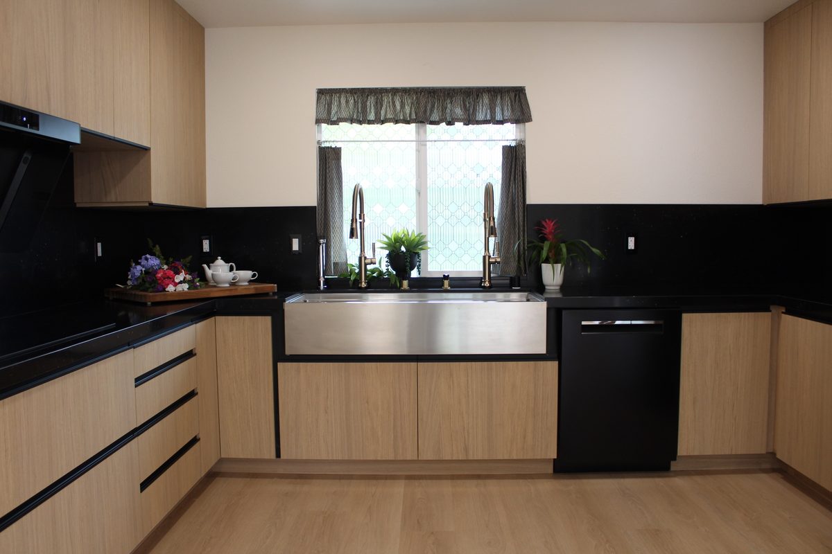 Straight-on view of the sink wall with light oak flat-panel cabinets, brass faucets, and black quartz backsplash