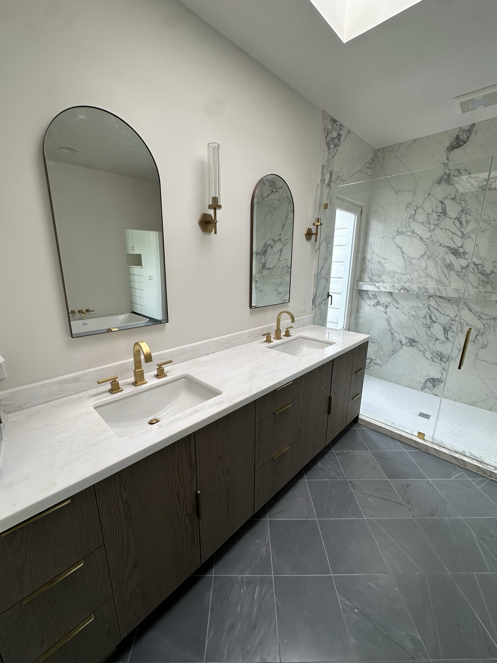 Primary bath vanity in dark stained wood with brass sconces, arched mirrors, and a marble shower beyond